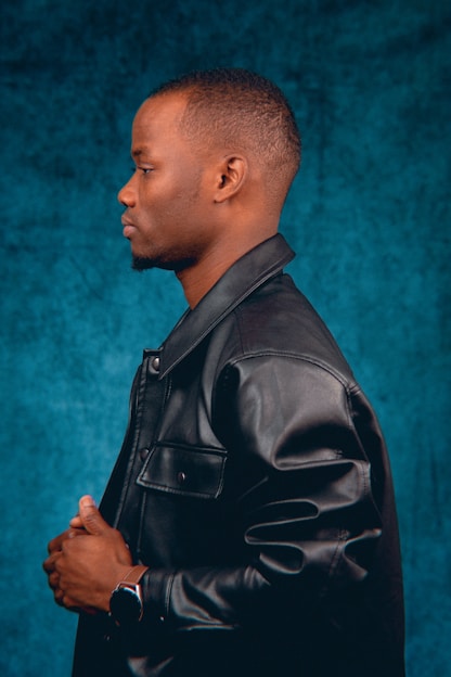 A person is shown in profile wearing a black leather jacket. The individual has short hair and is standing against a textured teal background, with hands slightly clasped in front. A watch is visible on the wrist.