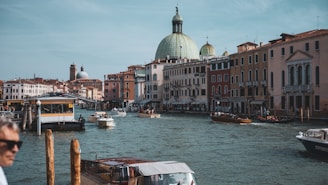 A scenic canal view in Venice with historical buildings lining the waterfront. Several boats are navigating the calm water, and a majestic domed building is prominently visible in the background. People can be seen in one of the boats, and a person is slightly blurred in the foreground.