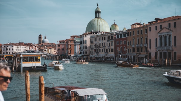 A scenic canal view in Venice with historical buildings lining the waterfront. Several boats are navigating the calm water, and a majestic domed building is prominently visible in the background. People can be seen in one of the boats, and a person is slightly blurred in the foreground.