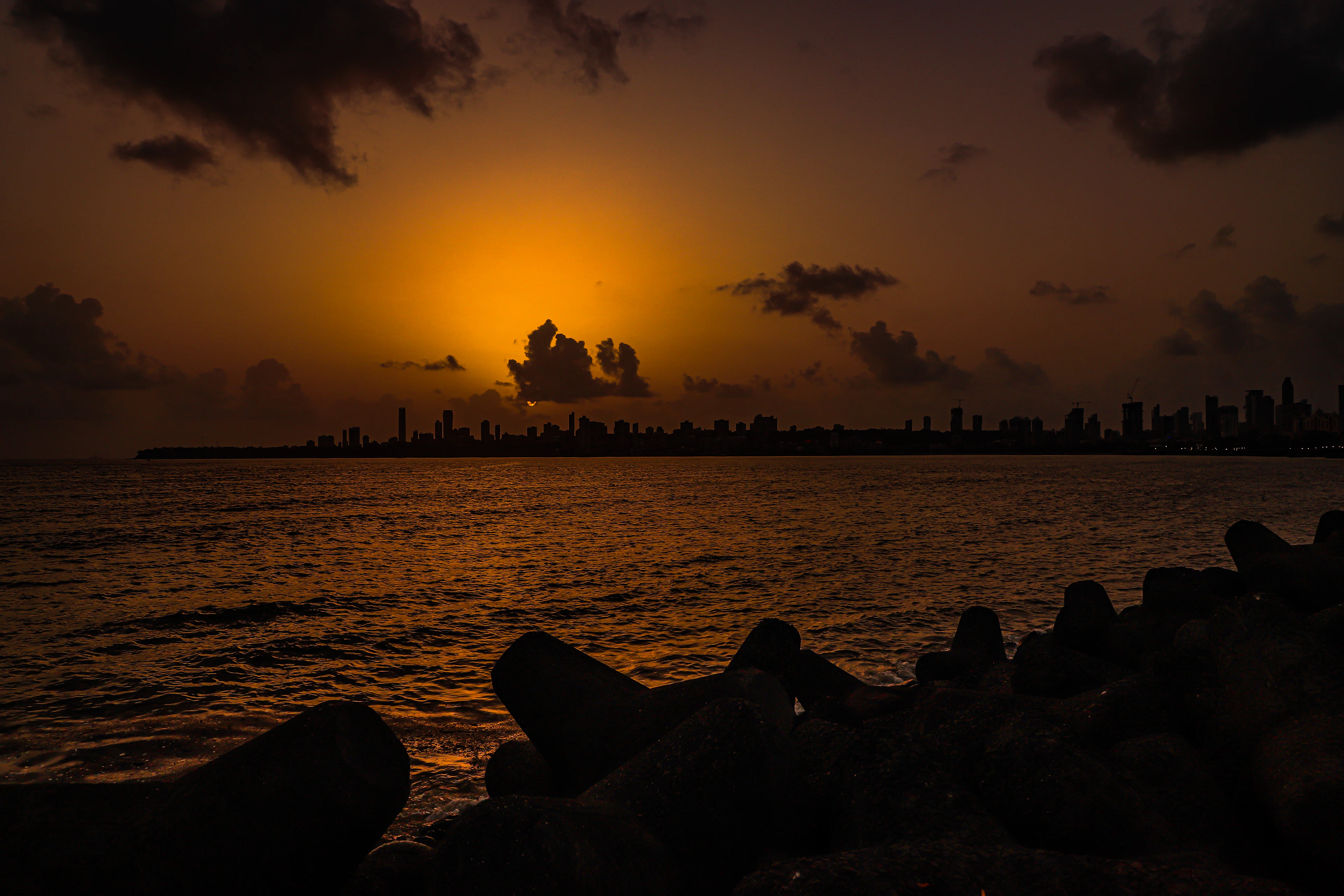 Sunset over the Mumbai skyline seen across the water from the Marine Drive tetrapods, the sun burning orange behind a low bank of cloud and the towers of the city reduced to silhouette.