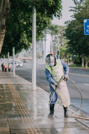 A person wearing protective gear, including a face shield and gloves, is power washing a sidewalk in an urban setting. Tall trees line the street, with a few people in the background walking along the sidewalk. The scene includes a road with light traffic and a prominent blue road sign.