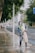 A person wearing protective gear, including a face shield and gloves, is power washing a sidewalk in an urban setting. Tall trees line the street, with a few people in the background walking along the sidewalk. The scene includes a road with light traffic and a prominent blue road sign.
