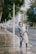 A person wearing protective gear, including a face shield and gloves, is power washing a sidewalk in an urban setting. Tall trees line the street, with a few people in the background walking along the sidewalk. The scene includes a road with light traffic and a prominent blue road sign.