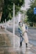 A person wearing protective gear, including a face shield and gloves, is power washing a sidewalk in an urban setting. Tall trees line the street, with a few people in the background walking along the sidewalk. The scene includes a road with light traffic and a prominent blue road sign.