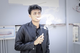 A person wearing a black leather jacket standing indoors, exhaling vapor from an electronic cigarette. The background includes a white electrical panel with circuit breakers and some visible wiring.