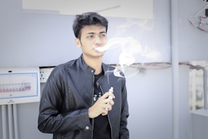 A person wearing a black leather jacket standing indoors, exhaling vapor from an electronic cigarette. The background includes a white electrical panel with circuit breakers and some visible wiring.