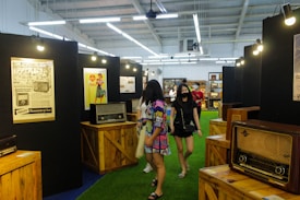 An indoor exhibition space features vintage radios displayed on wooden crates, surrounded by retro posters. People walk through the exhibition; two women in the foreground are looking around, one wearing a colorful dress and another in a black outfit with a mask.