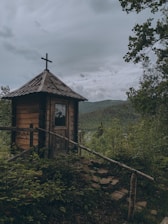 A small wooden chapel with a cross on top is situated amidst lush greenery, surrounded by trees and shrubbery. The chapel appears rustic and weathered, with a metal roof and wooden steps leading up to it. In the background, rolling hills and a cloudy sky create a serene and tranquil atmosphere.