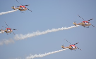 Four helicopters with colorful designs are flying in formation, emitting trails of white smoke against a clear blue sky.