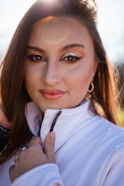 A close-up portrait of a person with long brown hair and striking makeup, including metallic eyeshadow and winged eyeliner. The person is wearing a white jacket with a zipper, holding it softly with one hand. The sunlight creates a halo effect around the head, adding a warm glow.
