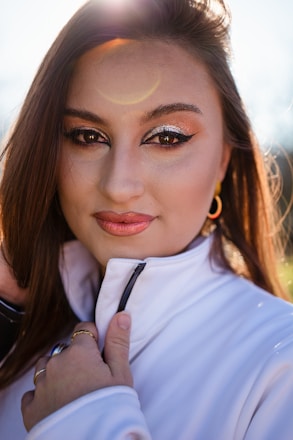 A close-up portrait of a person with long brown hair and striking makeup, including metallic eyeshadow and winged eyeliner. The person is wearing a white jacket with a zipper, holding it softly with one hand. The sunlight creates a halo effect around the head, adding a warm glow.