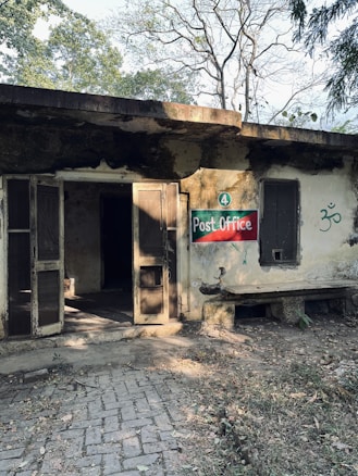 A weathered and aged building with an open doorway, surrounded by natural vegetation. The exterior shows signs of moss and grime accumulation. A hand-painted sign reading 'Post Office' is displayed on the wall. There is a simple stone bench beside the entrance and some scattered dry leaves on the paving stones in front.