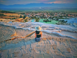 A person wearing a sun hat sits on the travertine terraces overlooking a stunning landscape with a vibrant town surrounded by greenery. The terraces are white and cascading, and the overall scenery includes expansive fields and a mountainous backdrop under a clear sky.