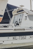 A side view of a white boat with the registration number KL-7352 visible on its body. The boat features a black steering wheel, a cabin with windows, and a radar on top. Part of another boat sails is seen in the background.