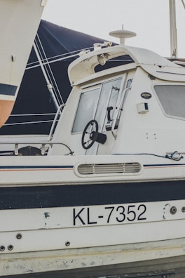 A side view of a white boat with the registration number KL-7352 visible on its body. The boat features a black steering wheel, a cabin with windows, and a radar on top. Part of another boat sails is seen in the background.