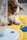 A dog intently focuses on a toy, which appears to be a blue ball. The dog, with a fluffy brown and white coat, is positioned on a bright yellow surface, with part of its face and paws visible. A blurred element in the foreground could be part of a dispenser or container.