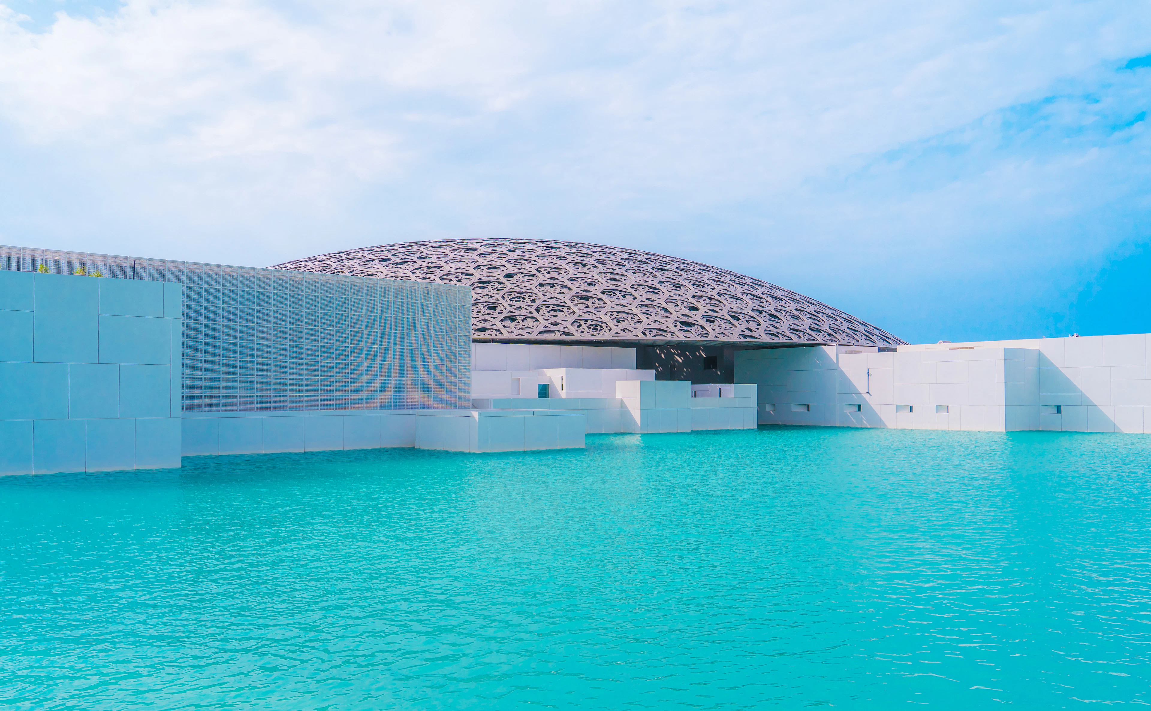The perforated steel dome of the Louvre Abu Dhabi resting over its white cubic galleries, the whole museum standing in flat turquoise seawater under a soft cloudy sky