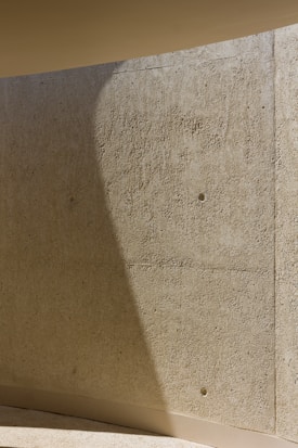 A textured concrete wall with a slightly curved surface. The wall casts a shadow that runs diagonally, adding depth to the scene. The light beige color of the concrete is highlighted by the overhead lighting.