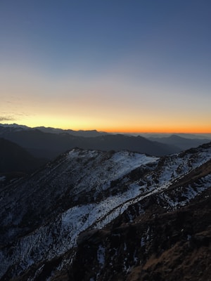 Snow-covered mountain peaks with rugged terrain illuminated by the warm hues of a setting or rising sun. The sky transitions from deep blues to vibrant oranges and yellows near the horizon, creating a dramatic and serene landscape.
