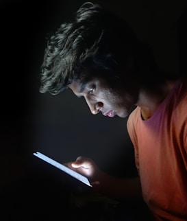 A person is intently looking at a glowing tablet in a dark setting, with the light illuminating their face and casting shadows.