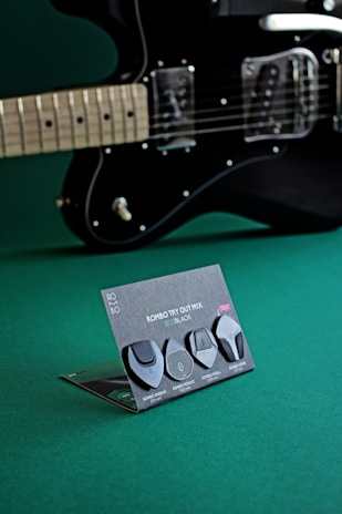 A black electric guitar is positioned with a focus on the strings and body. In front of the guitar, there is a display card featuring four different black guitar picks. Each pick is creatively named and described on the stand. The background is a solid green surface, providing contrast to the black guitar and picks.