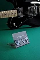 A black electric guitar is positioned with a focus on the strings and body. In front of the guitar, there is a display card featuring four different black guitar picks. Each pick is creatively named and described on the stand. The background is a solid green surface, providing contrast to the black guitar and picks.