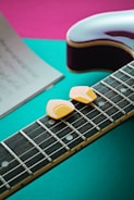 Two yellow guitar picks rest on the fretboard of an electric guitar. A sheet of music lies beside the guitar on a vibrant background combining teal and pink.