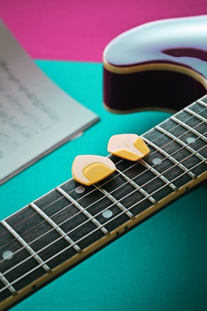 Two yellow guitar picks rest on the fretboard of an electric guitar. A sheet of music lies beside the guitar on a vibrant background combining teal and pink.