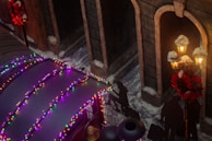 A street scene adorned with festive holiday lights in various colors on a structure or vehicle roof. Shadows of people walking with luggage are cast on the ground. Traditional lampposts decorated with red wreaths and snow provide a warm glow. The area is covered with patches of snow, enhancing the winter atmosphere.