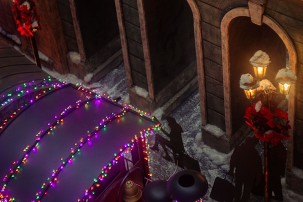 A street scene adorned with festive holiday lights in various colors on a structure or vehicle roof. Shadows of people walking with luggage are cast on the ground. Traditional lampposts decorated with red wreaths and snow provide a warm glow. The area is covered with patches of snow, enhancing the winter atmosphere.