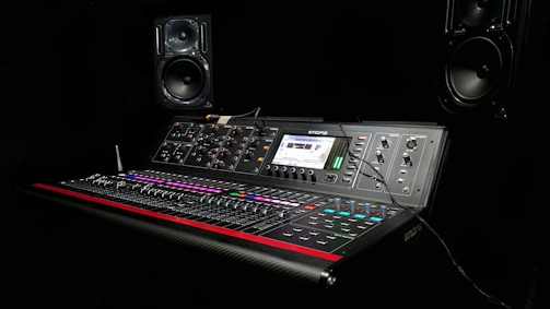 A professional audio mixing console is set against a dark background. The console features numerous sliders, knobs, and buttons with colorful lighting, and it is equipped with a digital display showing audio levels. Two large monitor speakers are positioned symmetrically on either side of the console.