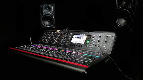 A professional audio mixing console is set against a dark background. The console features numerous sliders, knobs, and buttons with colorful lighting, and it is equipped with a digital display showing audio levels. Two large monitor speakers are positioned symmetrically on either side of the console.