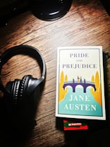 A set of black headphones rests on a wooden surface beside a copy of Jane Austen's 'Pride and Prejudice.' The book cover features an illustration of people standing on a bridge surrounded by tall trees in a contrasting palette of blue, yellow, and white. A small red sticker at the bottom indicates a bargain book sale.