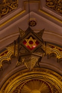 An ornate architectural detail featuring a large, hanging star-shaped light fixture with intricate designs and a prominent Star of David symbol in the center. The backdrop includes an arched ceiling with golden embellishments and patterns, creating a rich, lavish aesthetic.