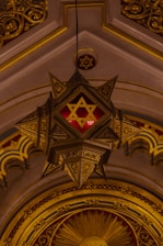 An ornate architectural detail featuring a large, hanging star-shaped light fixture with intricate designs and a prominent Star of David symbol in the center. The backdrop includes an arched ceiling with golden embellishments and patterns, creating a rich, lavish aesthetic.