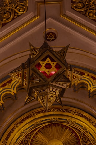 An ornate architectural detail featuring a large, hanging star-shaped light fixture with intricate designs and a prominent Star of David symbol in the center. The backdrop includes an arched ceiling with golden embellishments and patterns, creating a rich, lavish aesthetic.