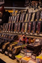 A variety of packaged goods are displayed on shelves in a market setting. The products are neatly arranged, with each shelf containing an assortment of items such as chocolate bars and possibly other gourmet products. Two people are visible in the background, adding a dynamic aspect to the scene. Bottles are placed on top of the display, enhancing the gourmet theme.