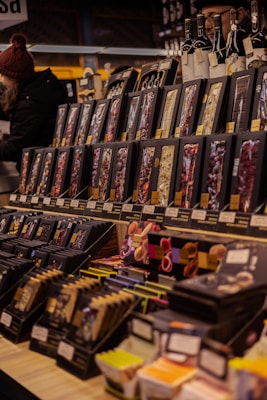 A variety of packaged goods are displayed on shelves in a market setting. The products are neatly arranged, with each shelf containing an assortment of items such as chocolate bars and possibly other gourmet products. Two people are visible in the background, adding a dynamic aspect to the scene. Bottles are placed on top of the display, enhancing the gourmet theme.