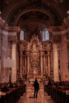 An ornate, richly decorated church interior featuring grand columns, intricate sculptures, and an elaborately carved altar. A person stands in the aisle with a tripod, suggesting photography or videography. People are seated in pews, indicating a gathering or service taking place.