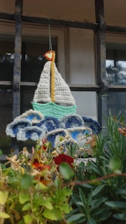A crocheted representation of a sailboat with a white sail and yellow mast hangs in front of a window with a metal grid. The sailboat is positioned above crocheted waves in hues of blue and white. Below the craftwork, various green plants and red flowers add a touch of nature.