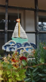 A crocheted representation of a sailboat with a white sail and yellow mast hangs in front of a window with a metal grid. The sailboat is positioned above crocheted waves in hues of blue and white. Below the craftwork, various green plants and red flowers add a touch of nature.