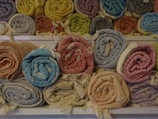 A collection of neatly rolled, colorful textiles is displayed on shelves. The fabrics have various patterns and colors, such as orange, blue, beige, pink, and yellow. They are arranged in two rows, with the upper row containing more fabrics.