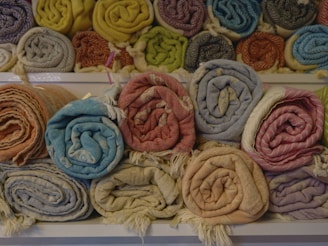 A collection of neatly rolled, colorful textiles is displayed on shelves. The fabrics have various patterns and colors, such as orange, blue, beige, pink, and yellow. They are arranged in two rows, with the upper row containing more fabrics.
