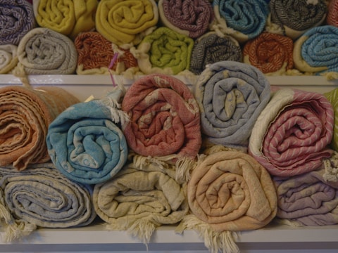 A collection of neatly rolled, colorful textiles is displayed on shelves. The fabrics have various patterns and colors, such as orange, blue, beige, pink, and yellow. They are arranged in two rows, with the upper row containing more fabrics.