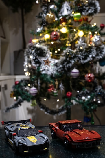 A decorated Christmas tree adorned with colorful ornaments and bright lights stands in the background. In the foreground, two model cars are displayed on a shiny surface. One is a black and yellow car with a spoiler, and the other is a red sports car.