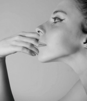 A black and white profile portrait of a person with a thoughtful expression. The person's hand is gently touching their lips and chin. The makeup includes prominent eyeliner, and their hair appears to be pulled back.