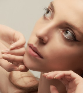 A close-up portrait of a person with subtle makeup, characterized by eyeliner accentuating the eyes. The face is framed by hands gently touching the chin and cheek. The expression appears contemplative, with eyes gazing upwards.
