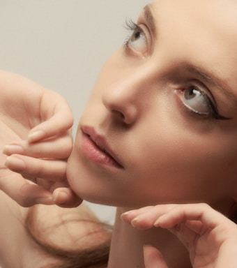 A close-up portrait of a person with subtle makeup, characterized by eyeliner accentuating the eyes. The face is framed by hands gently touching the chin and cheek. The expression appears contemplative, with eyes gazing upwards.