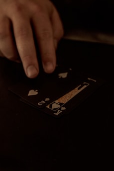 A hand is touching a stack of playing cards laid out on a dark surface. The cards are primarily black, featuring heart suits, including the king and jack. The scene is dimly lit, giving it an intimate or mysterious atmosphere.