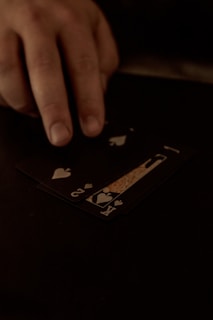 A hand is touching a stack of playing cards laid out on a dark surface. The cards are primarily black, featuring heart suits, including the king and jack. The scene is dimly lit, giving it an intimate or mysterious atmosphere.